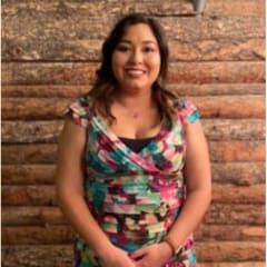 Mariah Ortiz, Marketing, in a floral dress in front of a log wall.